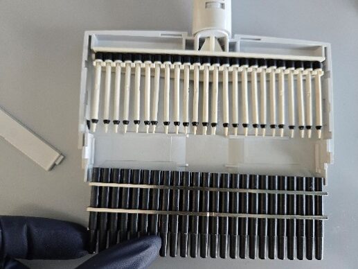 A gloved hand holds a gray multichannel plastic connector with black nose cones and white pistons arranged in rows on a light surface.