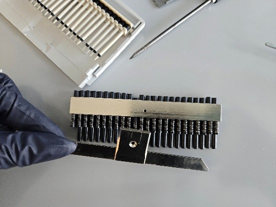 A gloved hand holds a metal part near a device with aligned nose cones and springs—during Nose Cone Replacement—on a gray surface, with tools and Eppendorf Research Plus components nearby.