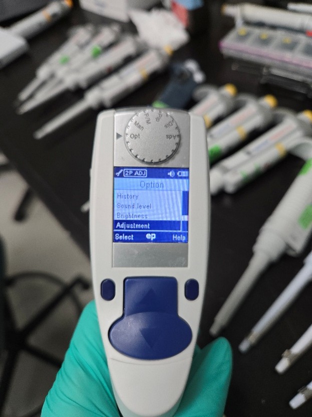A gloved hand holds an Eppendorf Xplorer digital pipette with a display screen showing adjustment instructions; several pipettes are visible on a lab bench in the background.