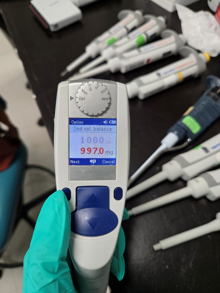 A gloved hand holds an Eppendorf Xplorer digital pipette displaying 2nd val. balance 1000 9970 mg in a lab setting, with other pipettes on the black workbench, ready for precise Eppendorf Xplorer Adjustment.