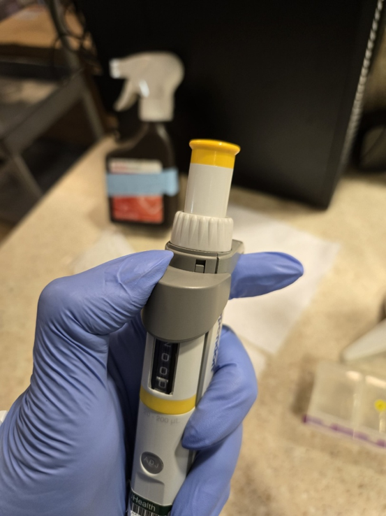 A gloved hand holds a Eppendorf Research Plus pipette with a yellow button, with lab equipment and a spray bottle blurred in the background.