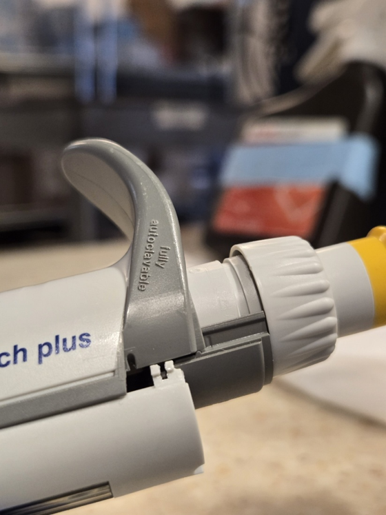 Close-up of a grey and yellow adjustable pipette on a lab bench with blurred laboratory equipment in the background, highlighting the importance of regular pipette calibration to ensure accurate measurements.