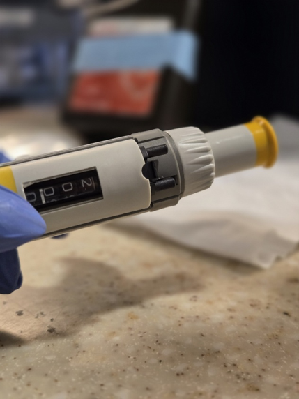 A gloved hand holding a laboratory micropipette set to a specific volume, with a blurred background of lab equipment—ensuring proper Eppendorf Research Plus calibration is essential to maintain accuracy and reliability in every experiment.
